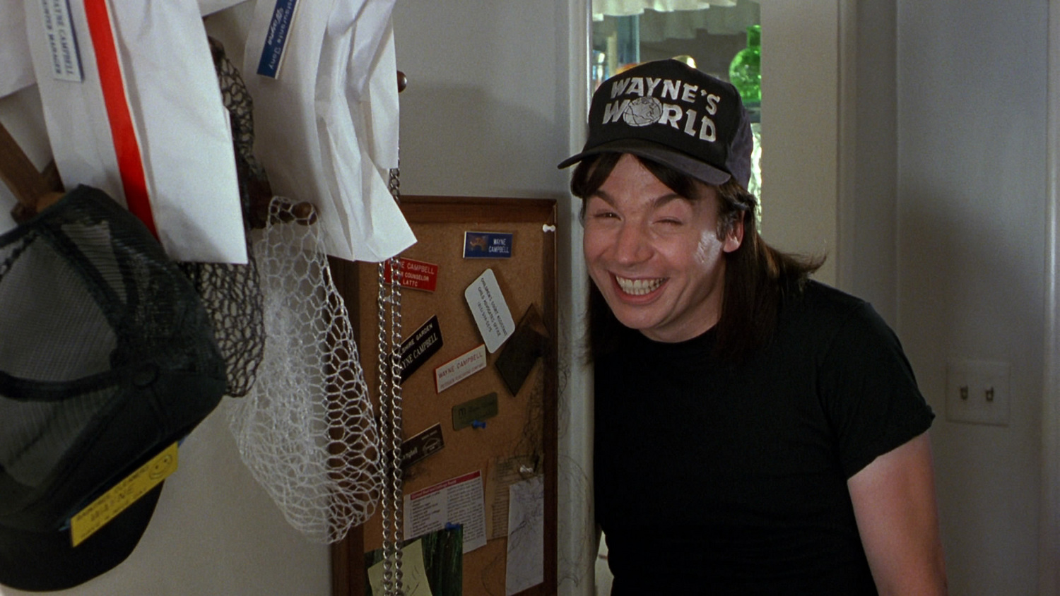 A screen grab of Wayne from the movie Waynes World, smiling awkwardly, next to his extensive collection of name tags and hair nets.